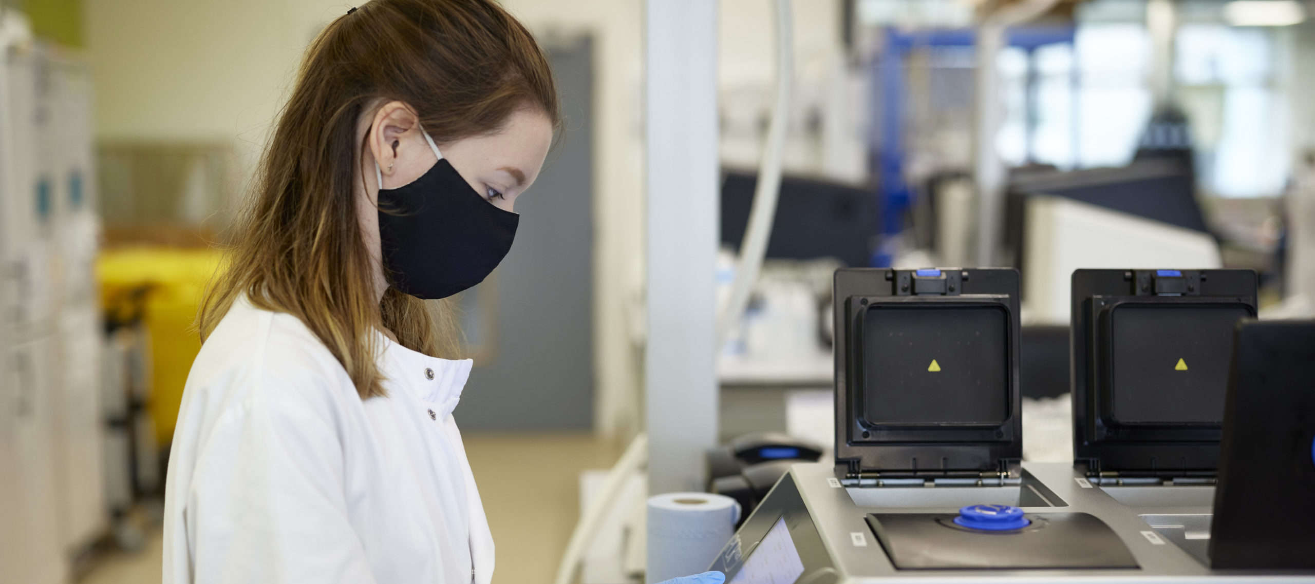 A woman in a laboratory wearing a face mask, focused on her work with scientific equipment around her.